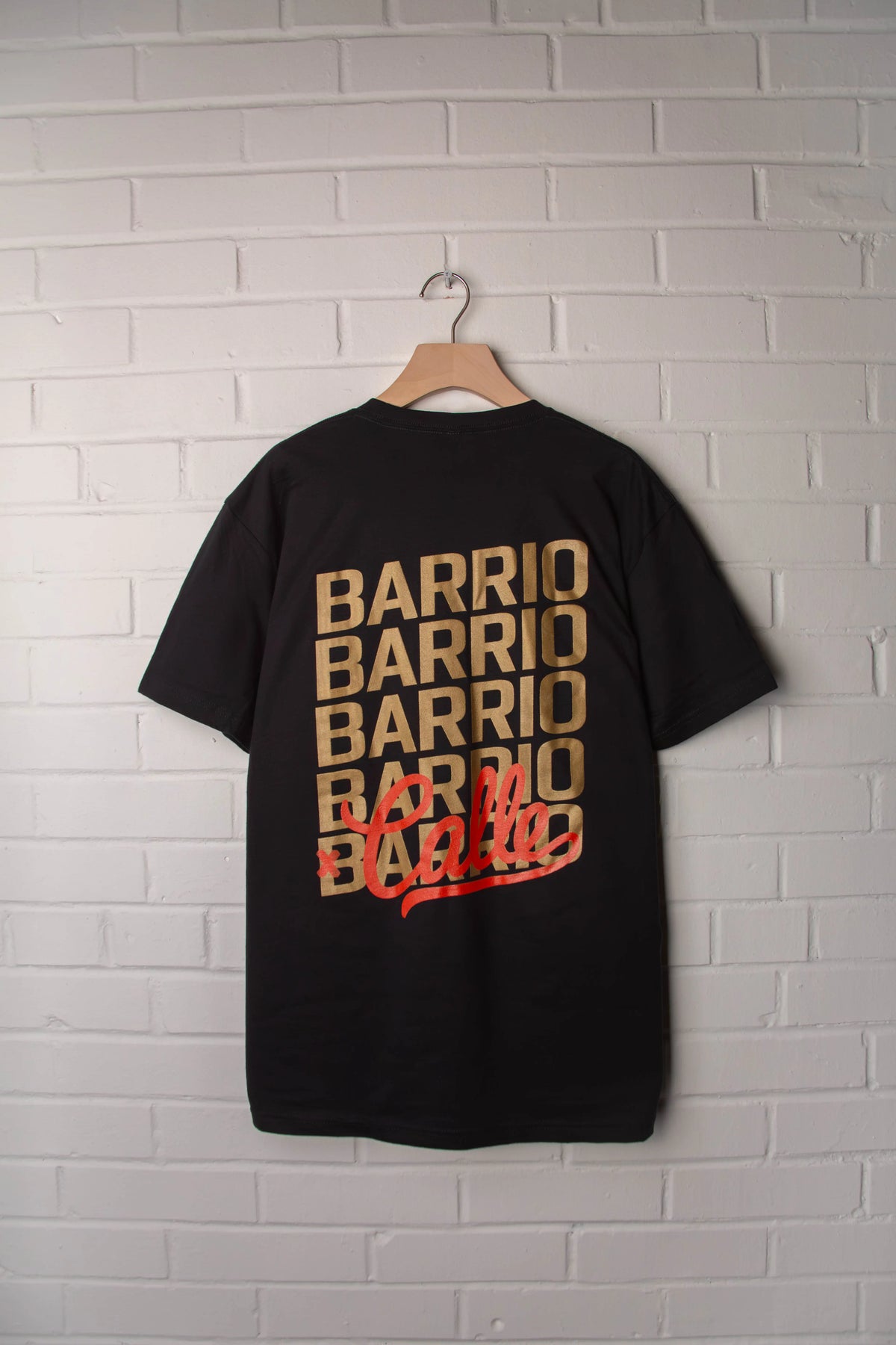 Black t-shirt with 'BARRIO' printed multiple times on a white brick wall background